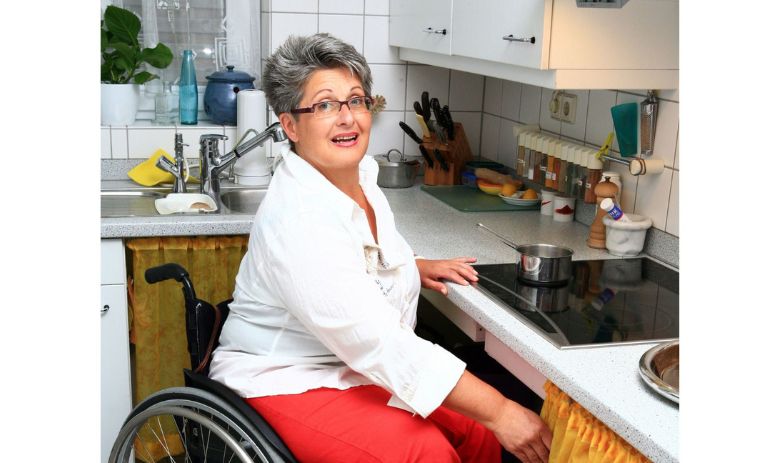  Une femme aux cheveux courts gris et portant des lunettes est assise dans un fauteuil roulant devant la cuisinière dans sa cuisine.