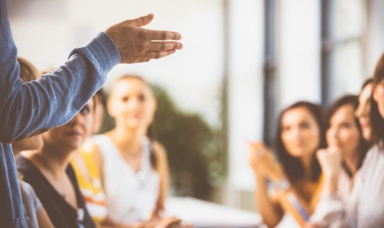 Unscharfes Bild von mehreren Personen an einem Tisch, die eine Person im Vordergrund anschauen. Von dieser ist nur der Arm zu sehen, sie macht eine Geste mit der Hand.