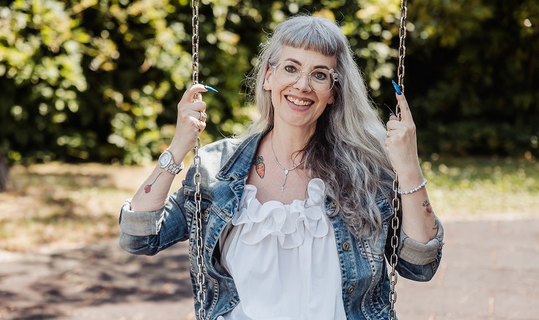 Chloé Buchmann Sanroma, eine modische Frau mit langen grauen Haaren sitzt auf einer Schaukel und lächelt in die Kamera.